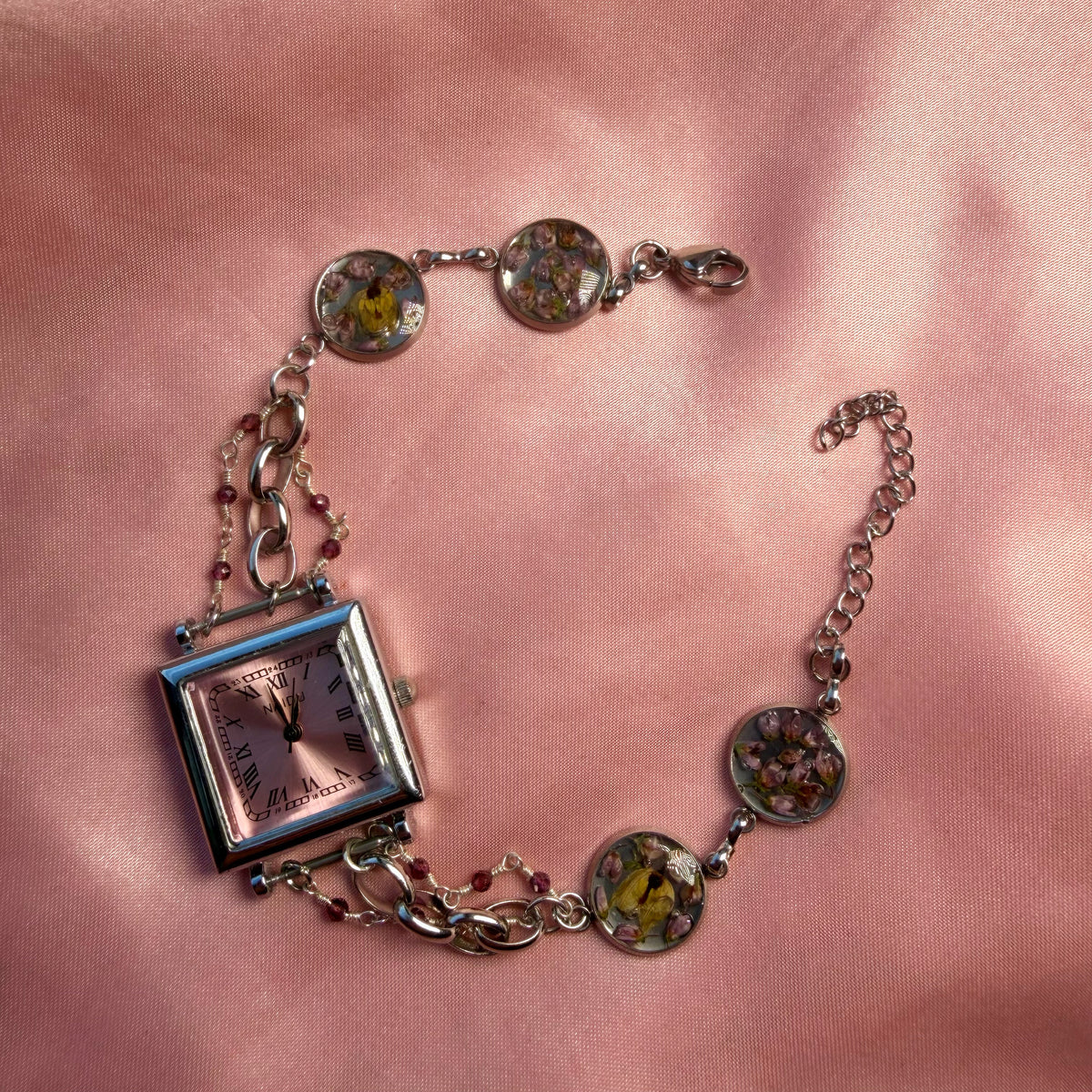 A silver-tone bracelet with a square pink dial and a handmade floral band